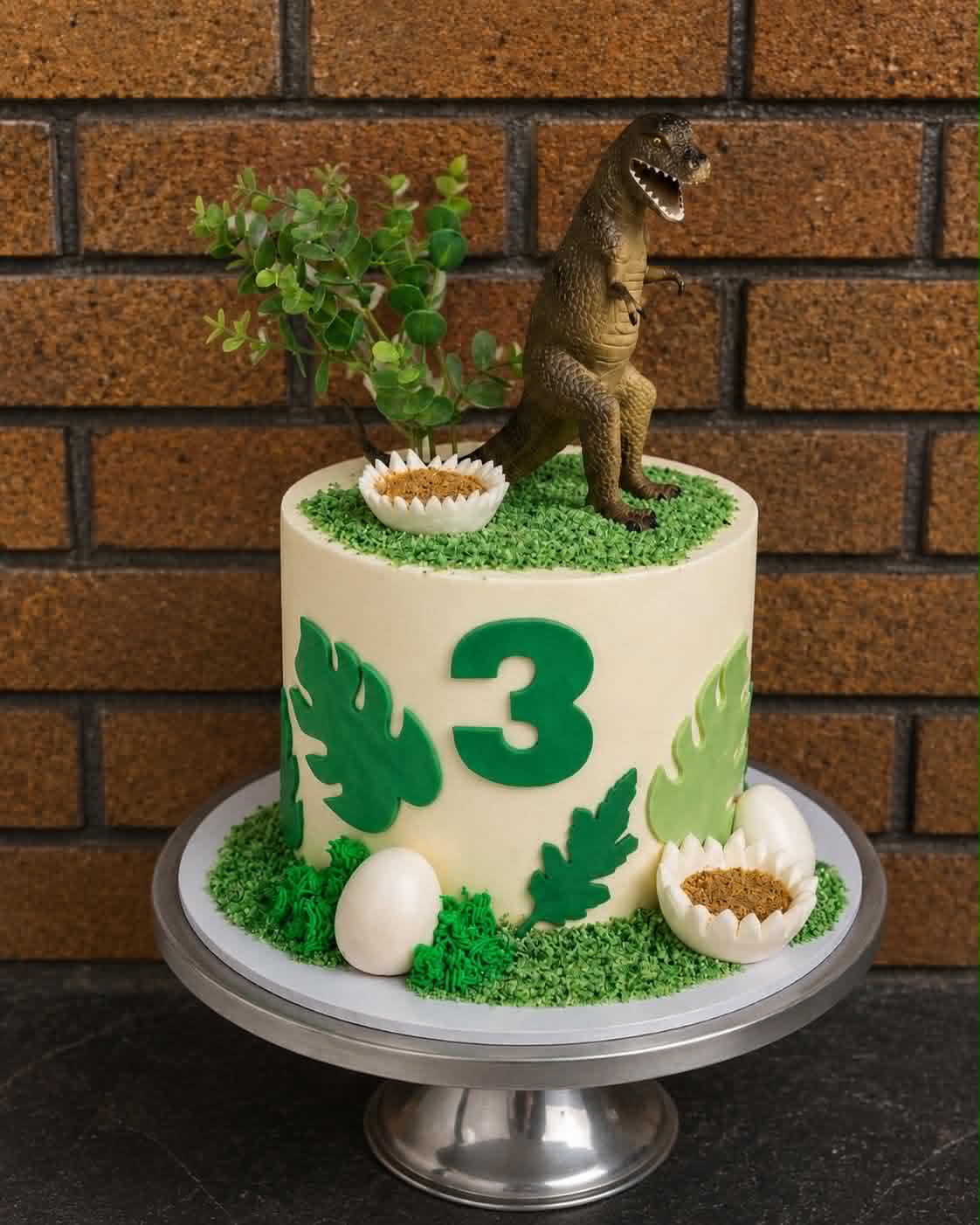 Dinosaur 3rd birthday cake with T-Rex topper, green grass, monstera leaves and dinosaur eggs